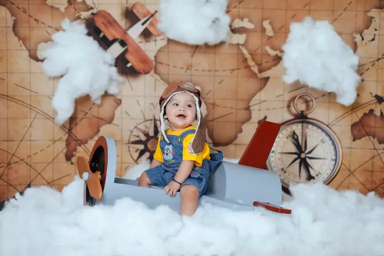 Baby in a pilot cap and goggles on a toy aeroplane among cotton-wool clouds in a world-map set