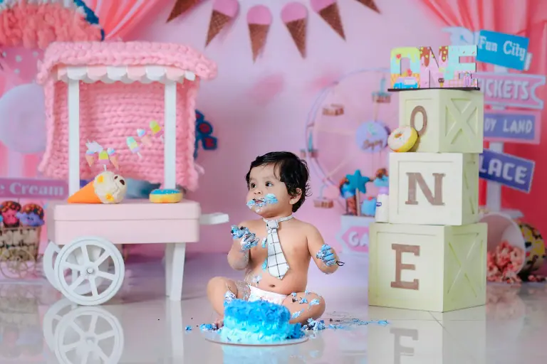 Baby with blue cake on face and hands beside a pink ice-cream cart and ONE letter blocks